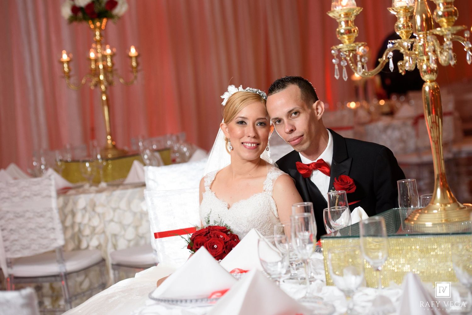Smiling bride and groom seated at decorated wedding reception table with gold candelabras and red roses.