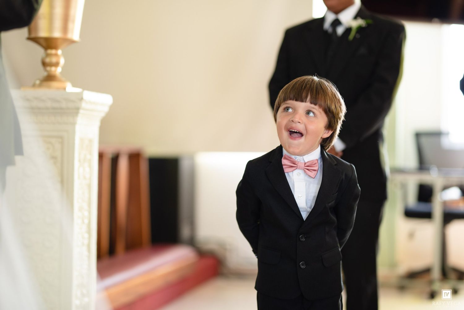 Young ring bearer in black suit and pink bow tie looks up with excitement at a wedding ceremony.