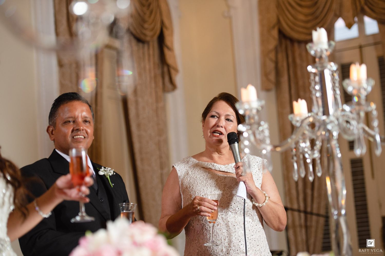 Woman speaks into microphone at elegant wedding reception while man holds champagne glass beside crystal candelabra.
