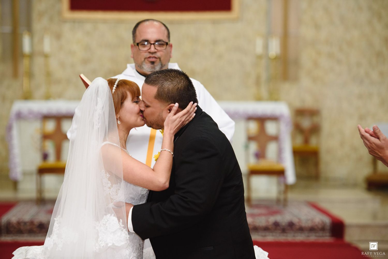 Newlyweds share first kiss at church altar as officiant watches during wedding ceremony.