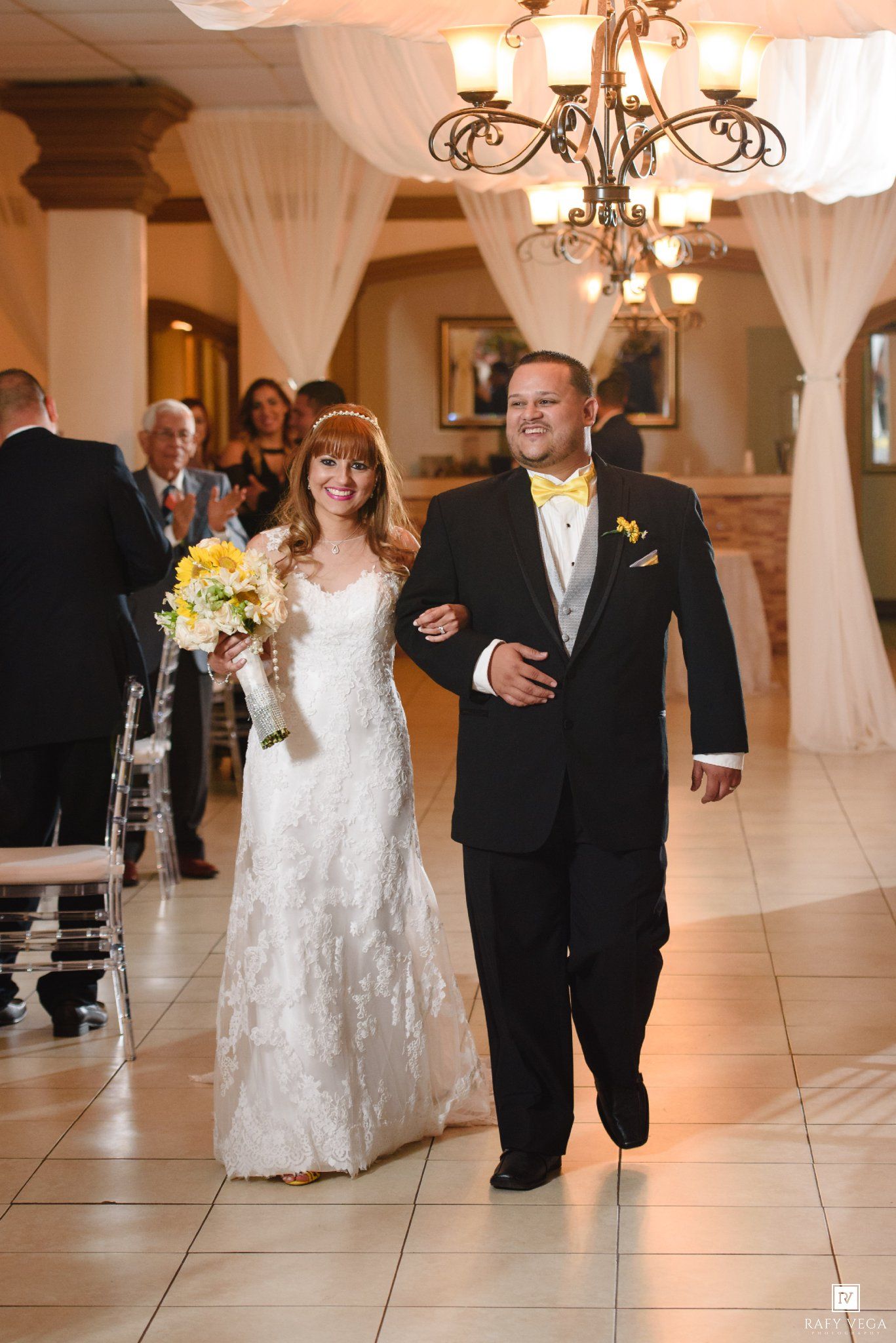 Bride in white lace gown and groom in black tuxedo with yellow bow tie walk down wedding reception aisle.
