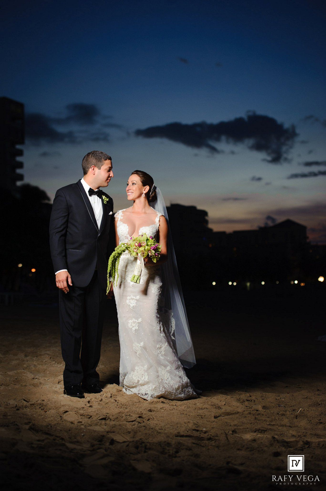 Bride and groom share a romantic moment on a sandy beach at sunset, bride in lace gown holding floral bouquet.