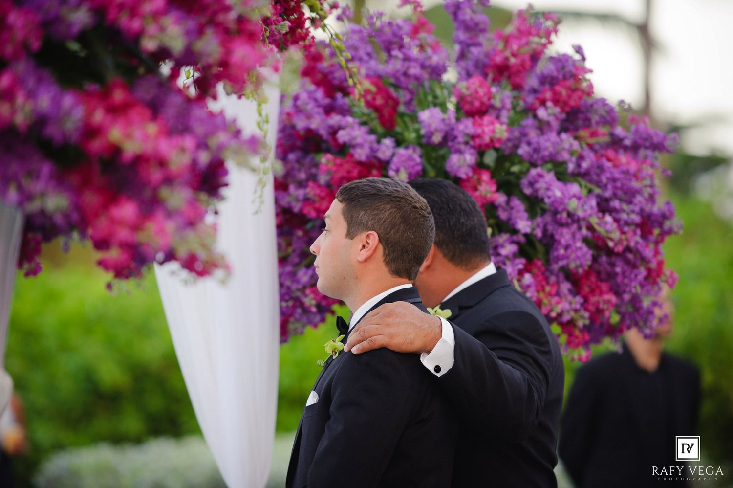 Two grooms embrace tenderly at outdoor wedding ceremony beneath stunning purple and pink floral arch.