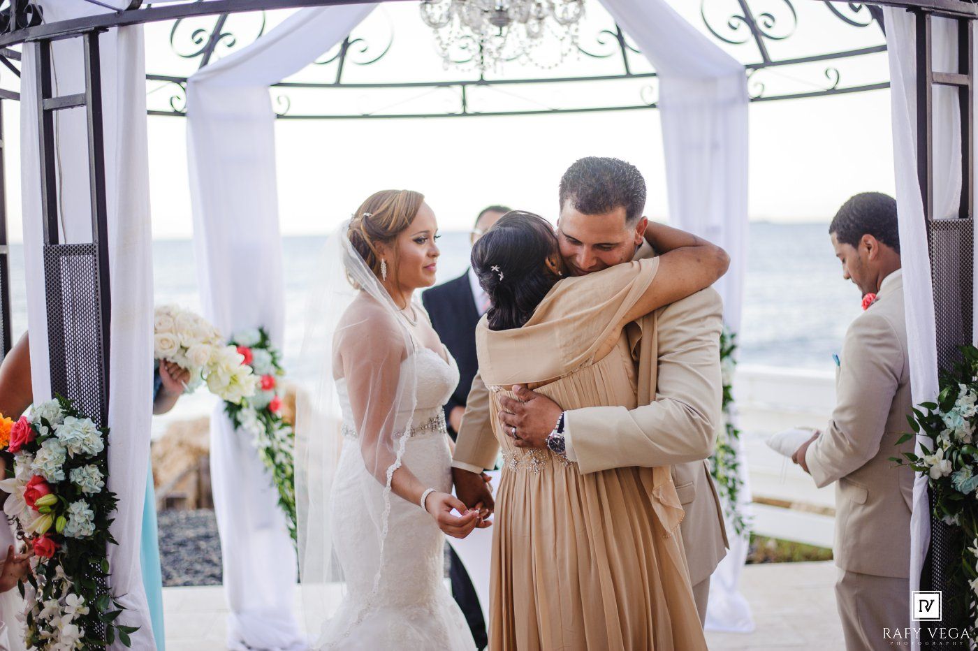 Bride and groom share emotional hug with guest during outdoor beach wedding ceremony under white floral arch.