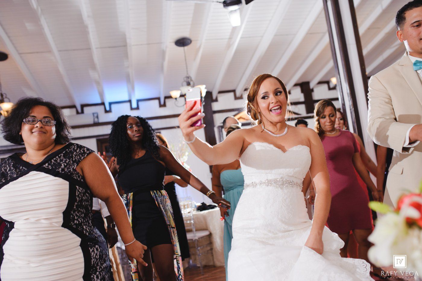 Bride in white gown takes selfie with guests during lively wedding reception celebration.