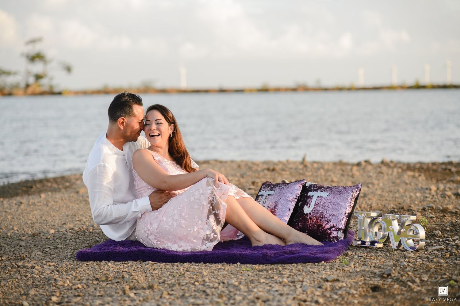 Couple sitting on purple mat at rocky beach shore, woman laughing joyfully as man embraces her at sunset.
