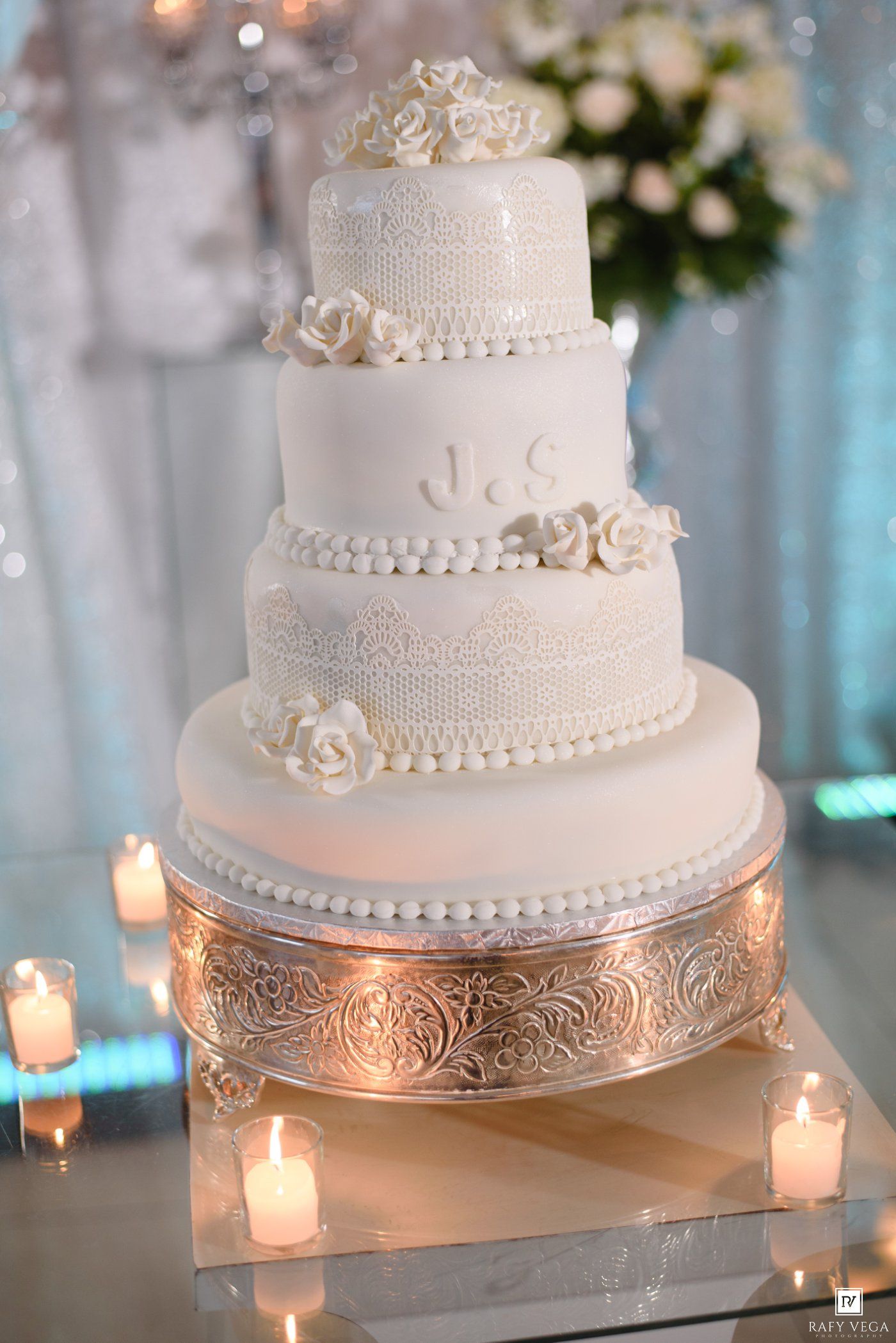 Elegant four-tier white wedding cake with lace details, pearl borders, sugar roses, and monogram initials on a gold stand.