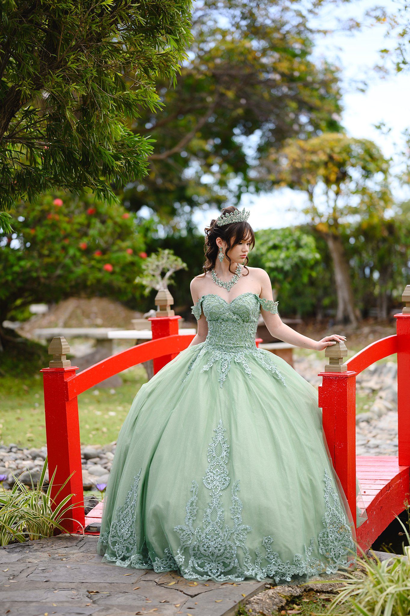 Young woman in mint green ball gown with silver embroidery and crown poses on red bridge in lush garden.