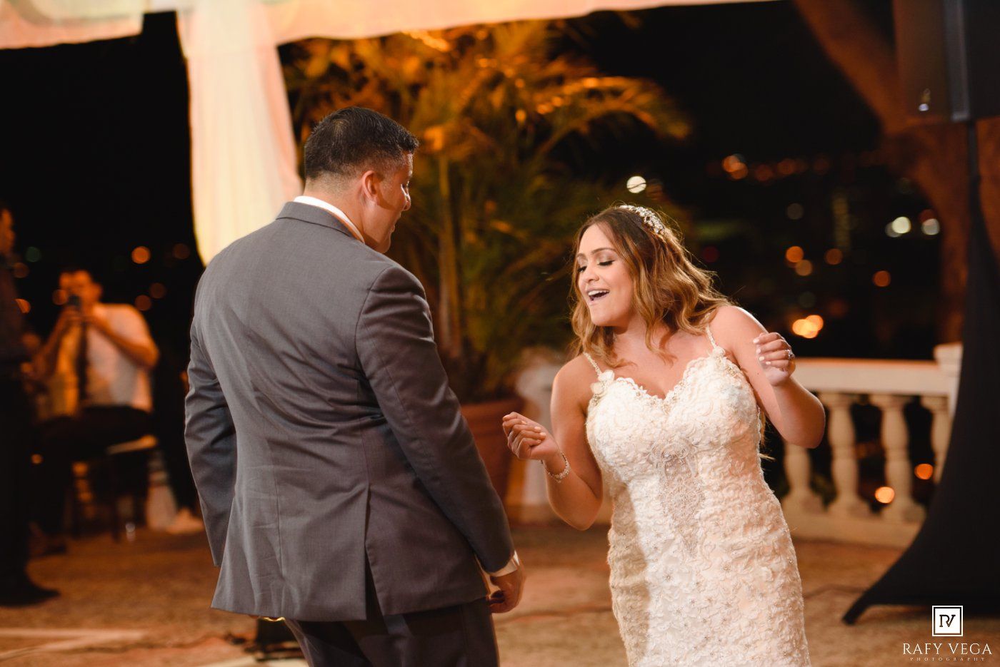 Bride in lace mermaid gown laughs joyfully while dancing with groom outdoors at romantic evening wedding reception.