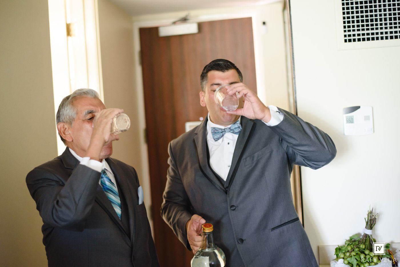 Two men in suits drinking shots together at a wedding celebration indoors.