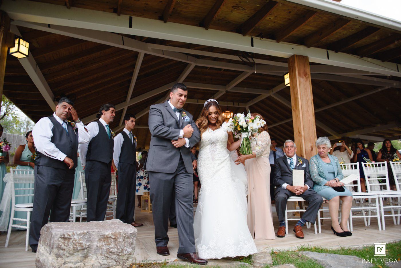 Bride and groom walk down the aisle together during an outdoor wedding ceremony under a wooden pavilion.
