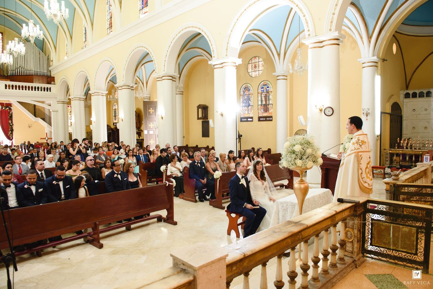 Elegant church wedding ceremony with guests seated in wooden pews as couple exchanges vows at ornate altar.