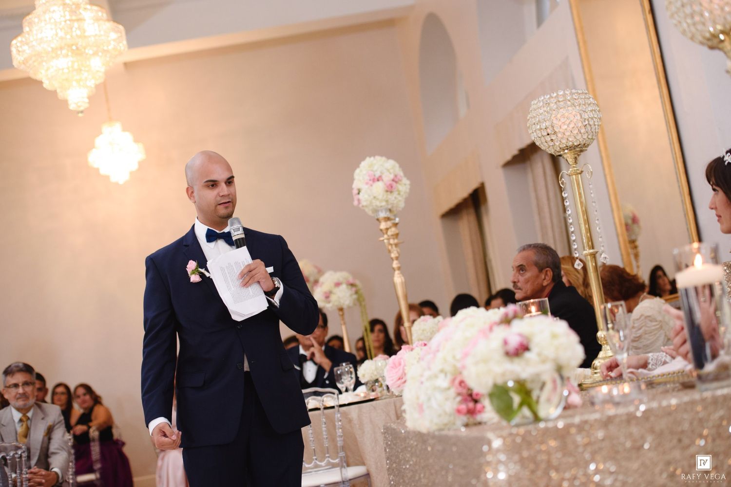 A bald groom in a navy tuxedo gives a speech at an elegant wedding reception with floral centerpieces.