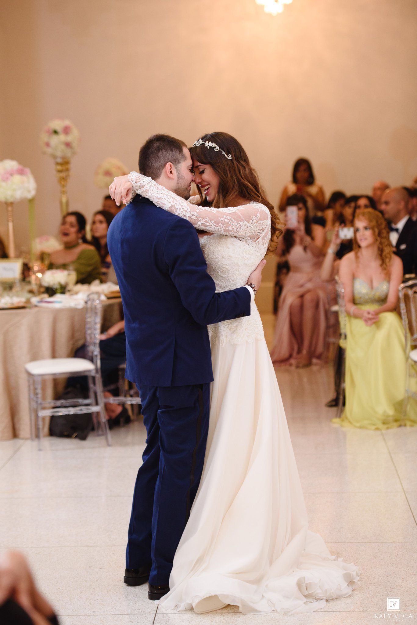 Bride and groom sharing their first dance at wedding reception, guests watching in background, elegant ballroom setting.