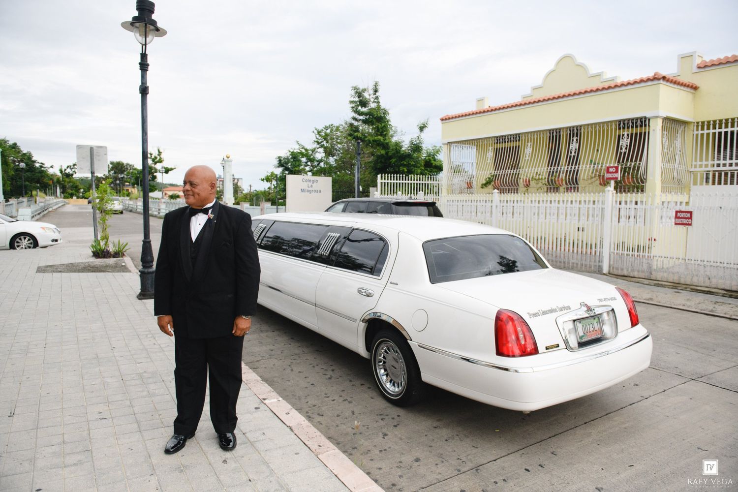 Man in tuxedo standing beside a white stretch limousine on a city street near a pink building.