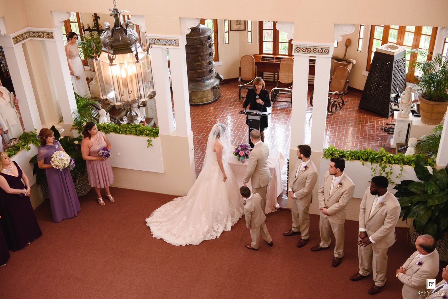 Aerial view of an elegant indoor wedding ceremony with bride in white gown, groomsmen in tan suits, and bridesmaids in purple dresses.
