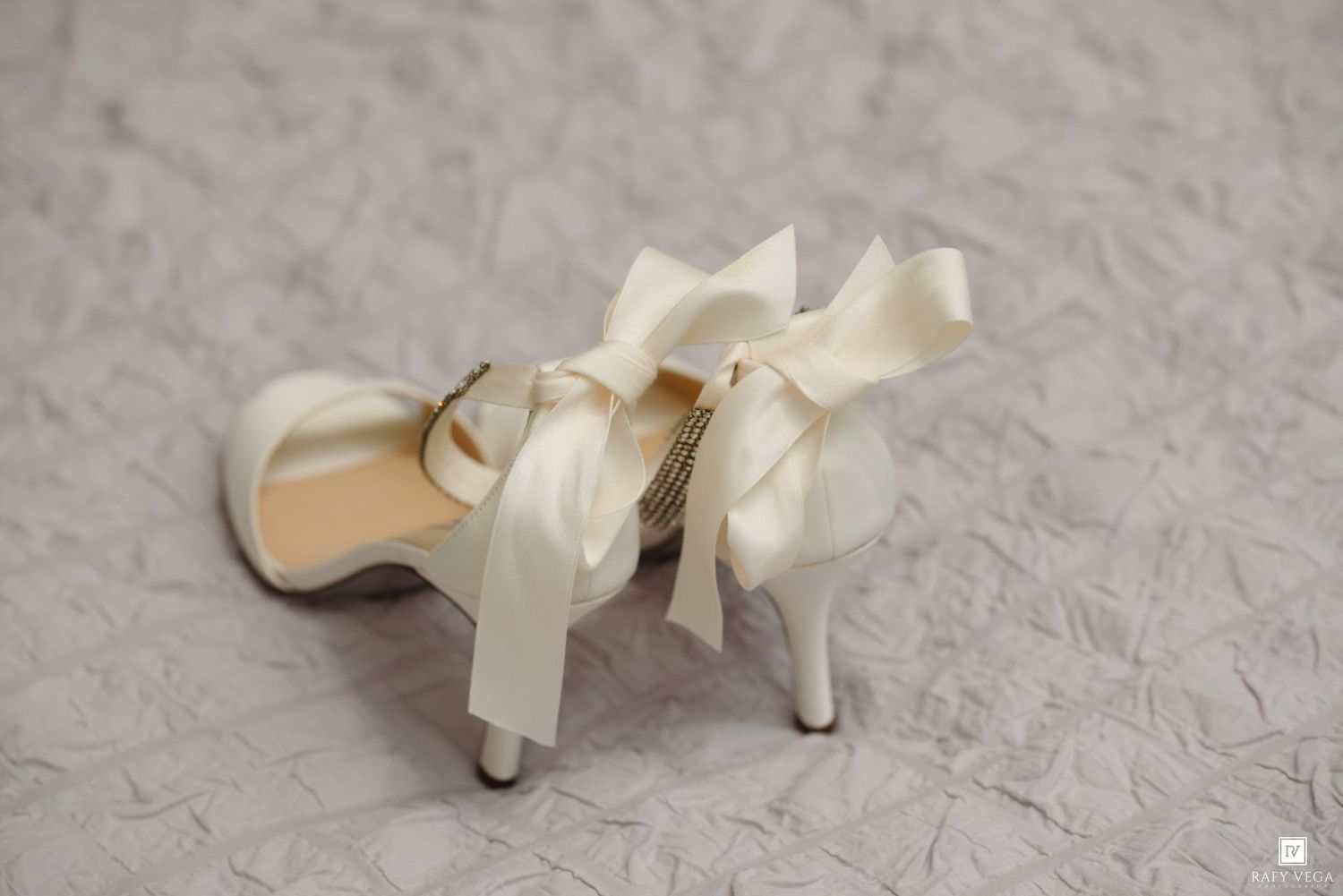 Elegant ivory satin bridal heels with large ribbon bows, displayed on a white lace background.