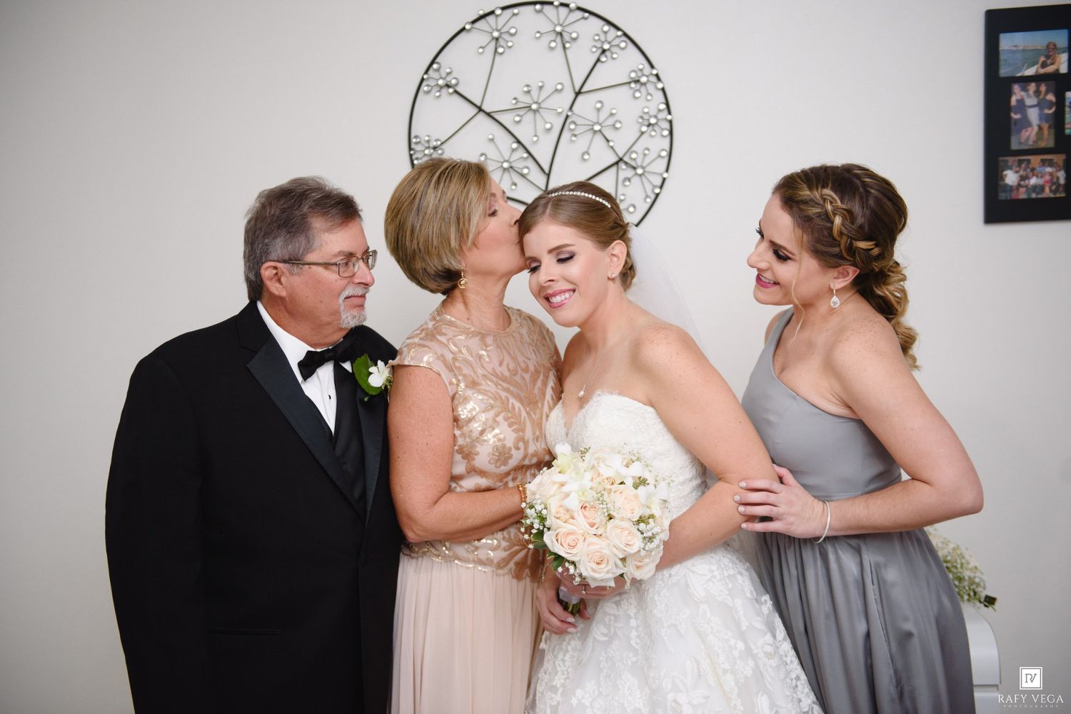 A bride in a white gown holds a bouquet while posing with her family, including parents and a bridesmaid, on her wedding day.