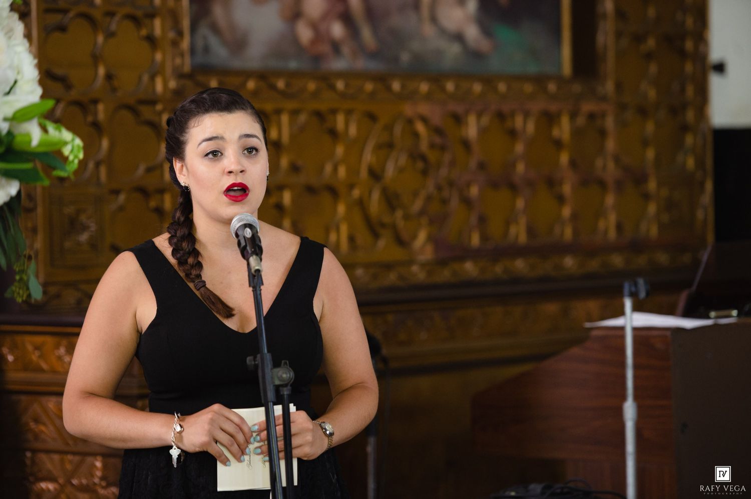 Woman in black dress singing at microphone in ornate venue with golden decor.