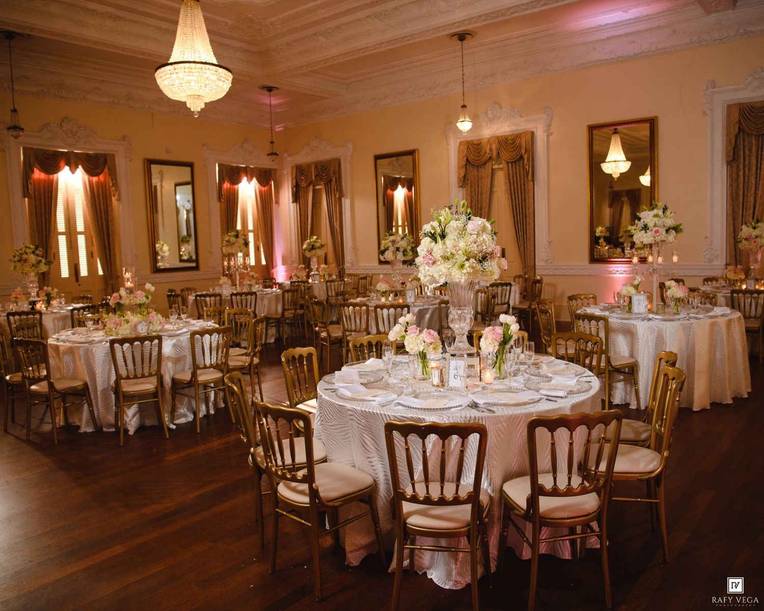 Elegant wedding reception hall with round tables, gold chairs, white floral centerpieces, and warm chandelier lighting.
