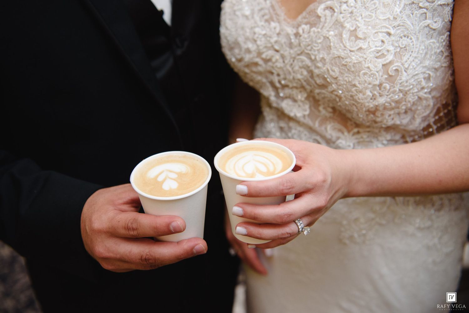 Bride and groom hold matching latte art coffees in white cups at their wedding.