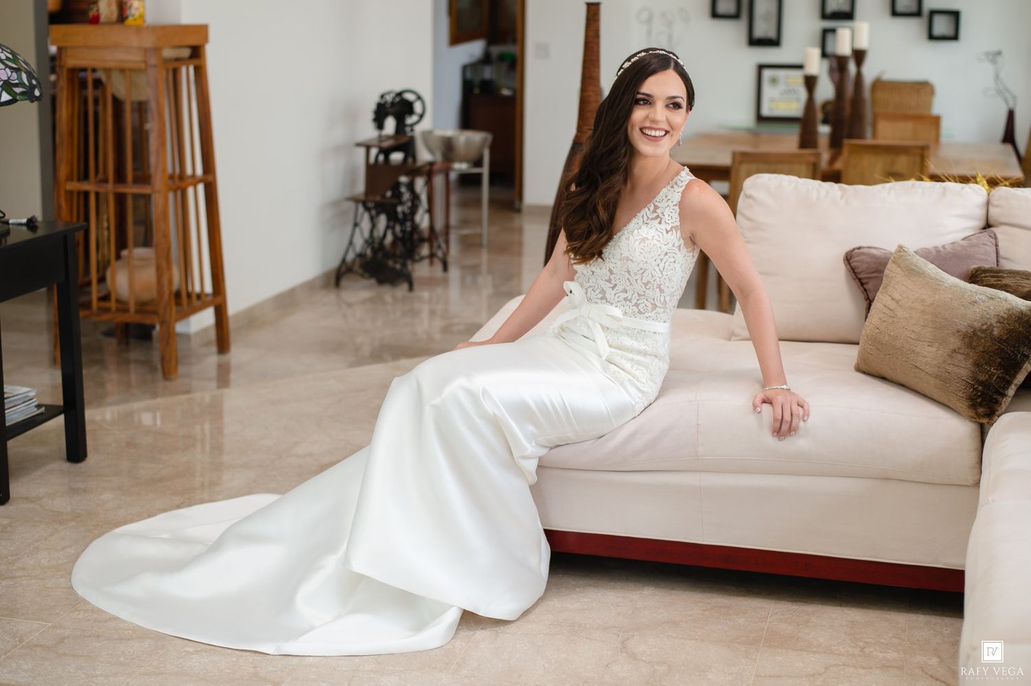 Bride in a white lace wedding gown with a long train sits elegantly on a beige sofa, smiling and wearing a tiara.