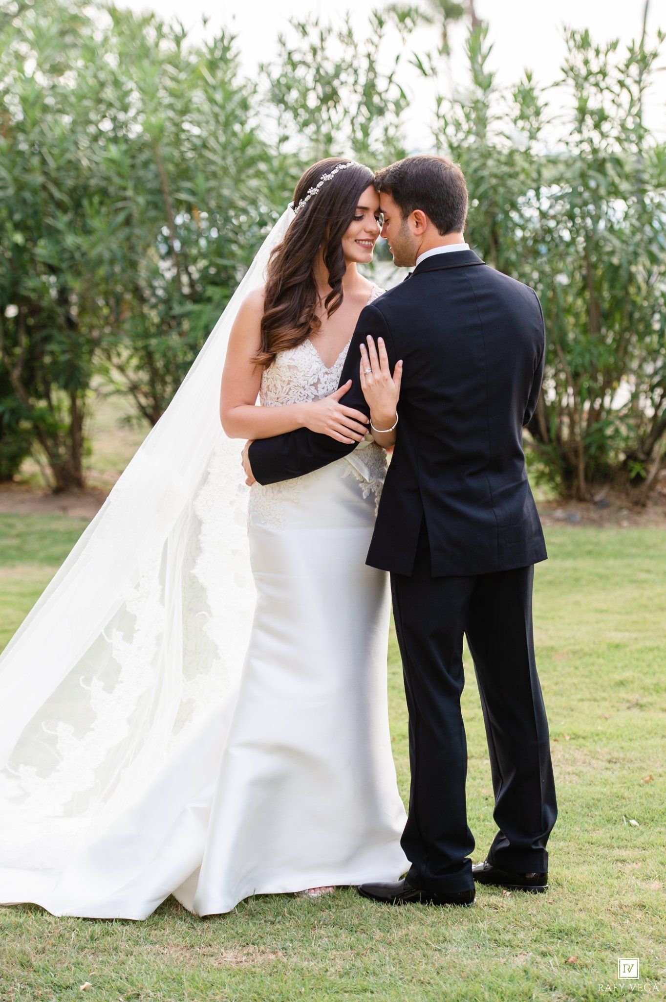 Bride in lace wedding gown with cathedral veil sharing romantic moment with groom outdoors.