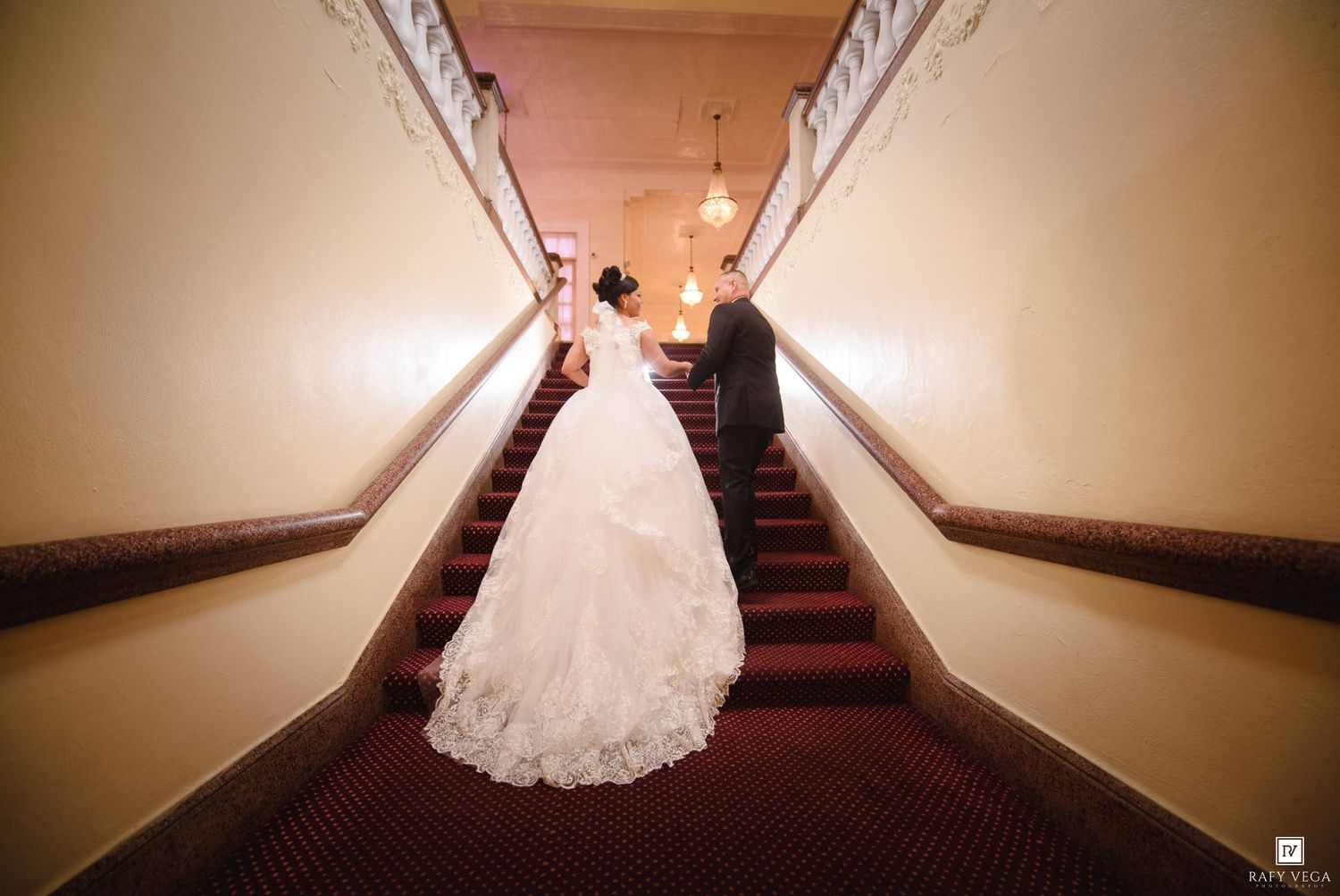 Bride and groom ascending elegant staircase with red carpet, bride in flowing white gown at wedding venue.