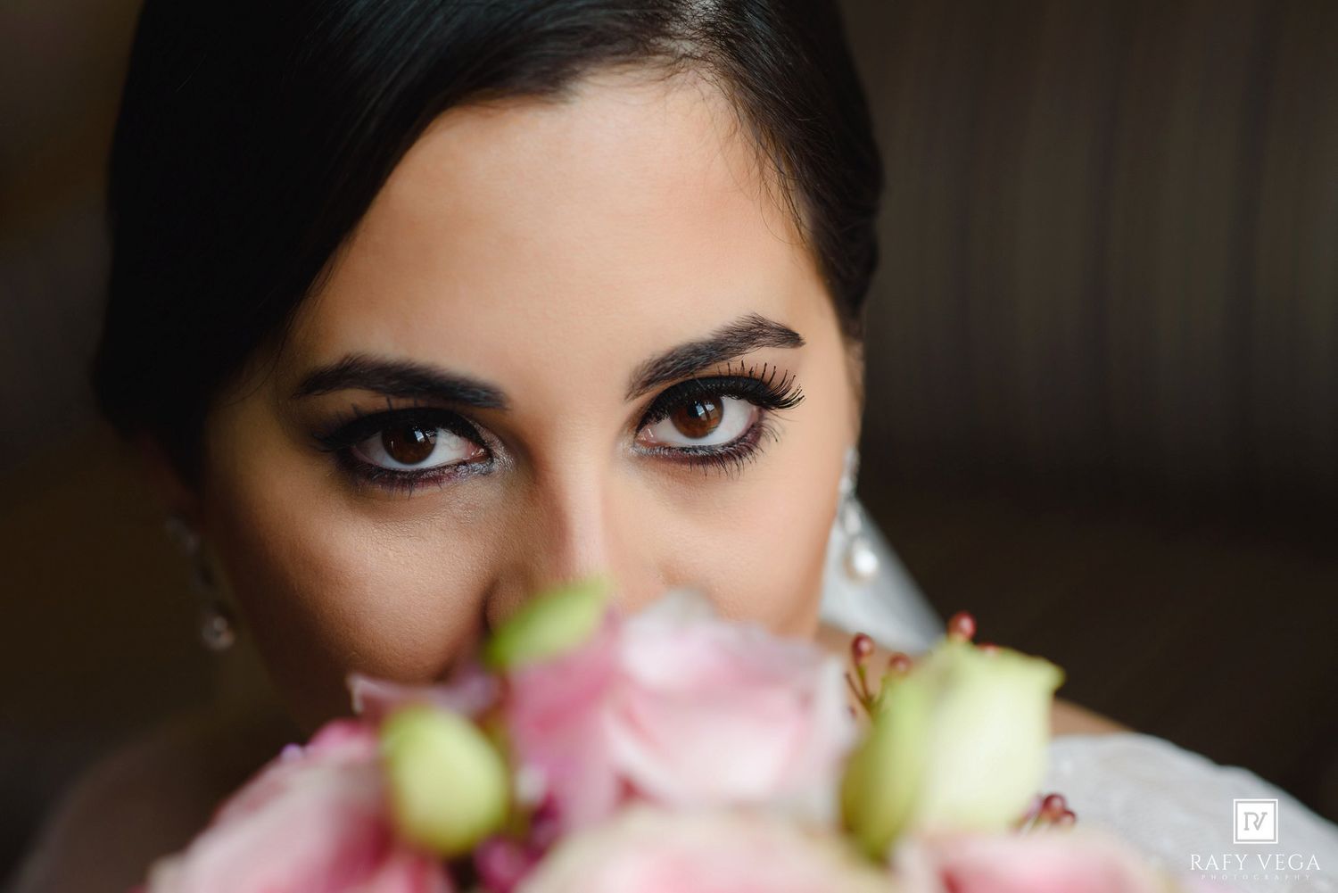 Close-up of a bride with smoky eye makeup holding a bouquet of pink and white roses near her face.