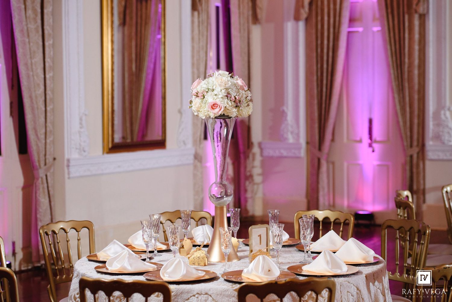 Elegant wedding reception table with tall floral centerpiece, gold chairs, and pink uplighting in a ballroom.
