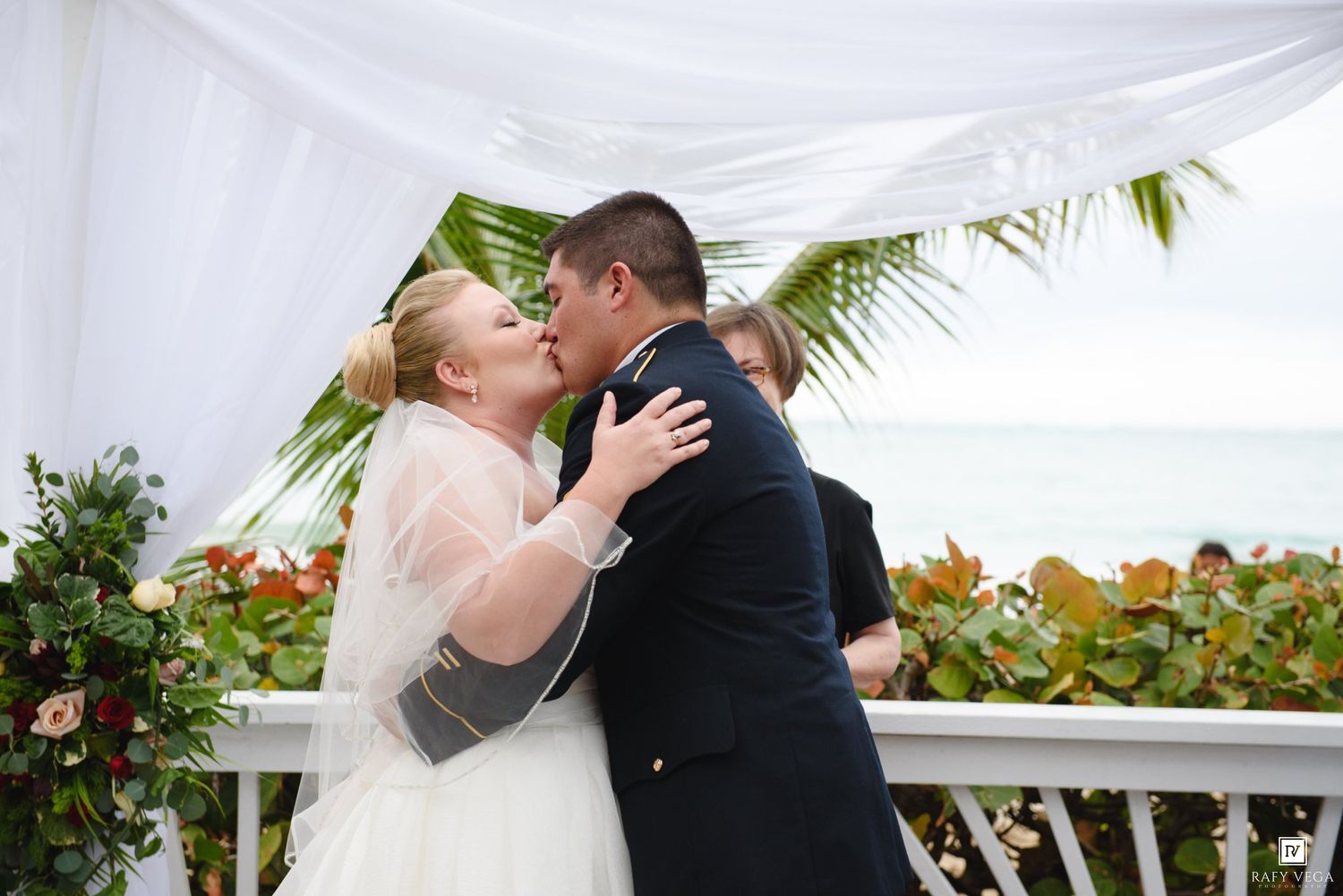 Bride and groom share their first kiss at an outdoor beach wedding ceremony under a white draped arch.