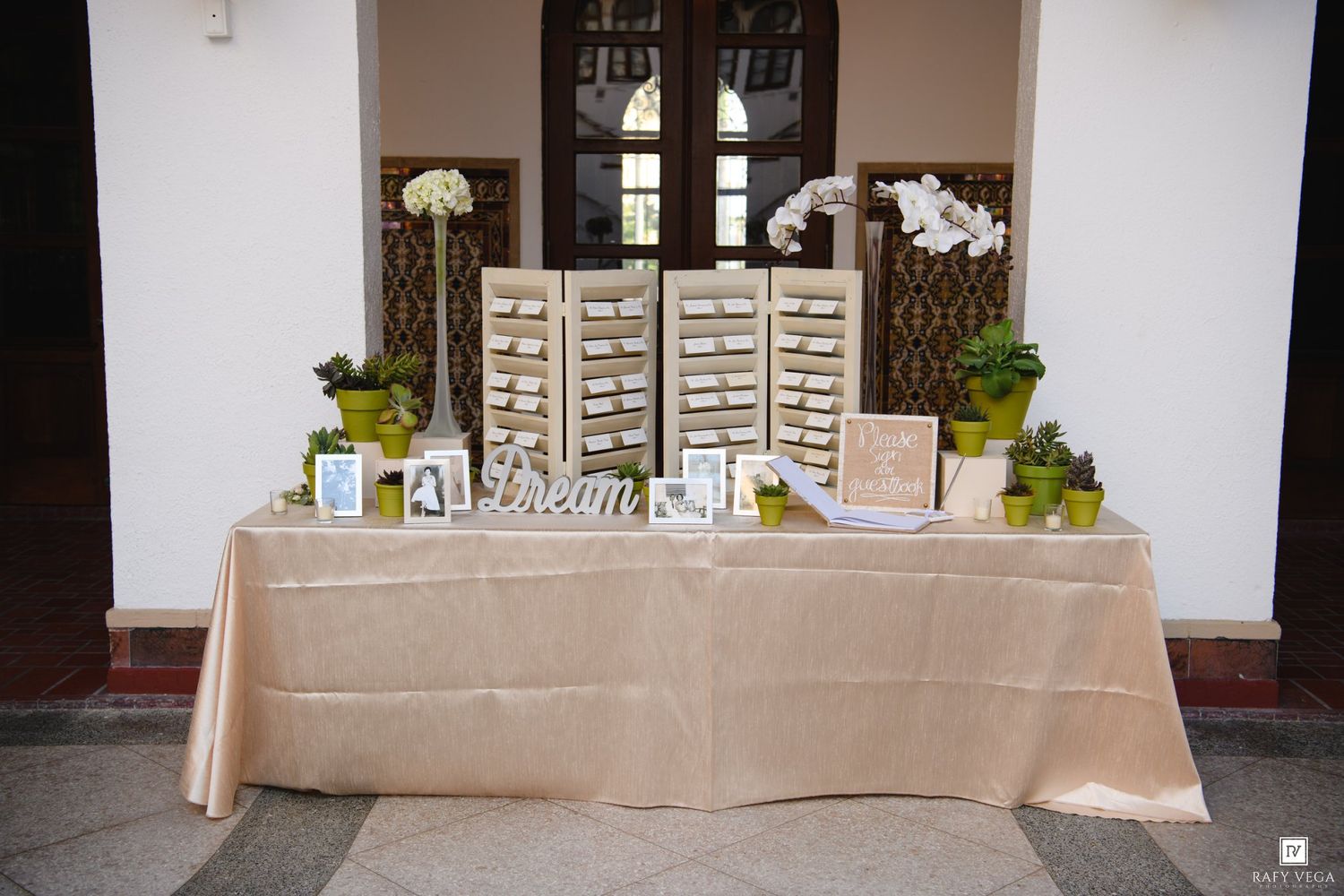 Elegant wedding seating chart display on a gold tablecloth with white shutters, orchids, and green plants at a venue.