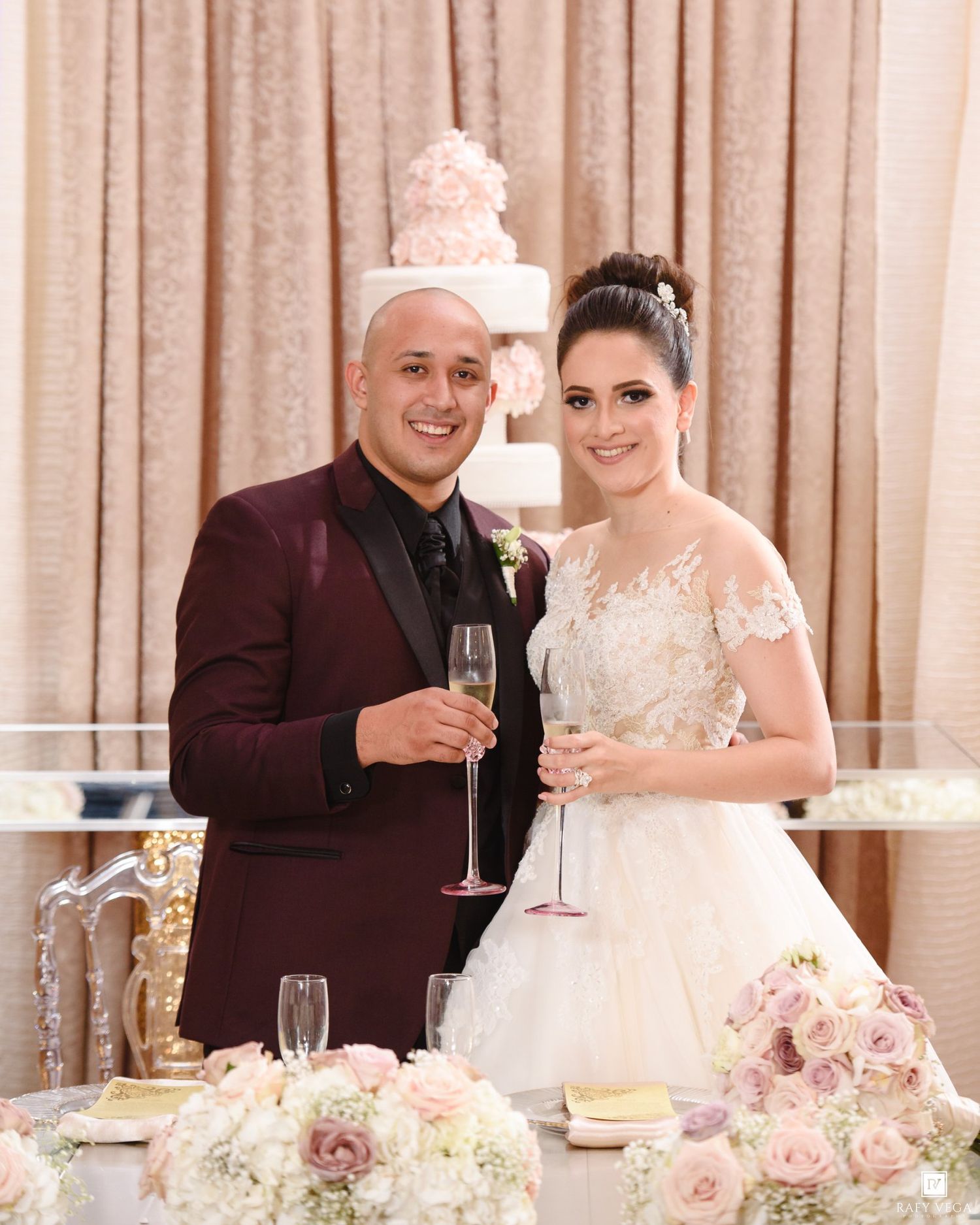 Bride and groom toasting with champagne at wedding reception table with floral arrangements and tiered cake.