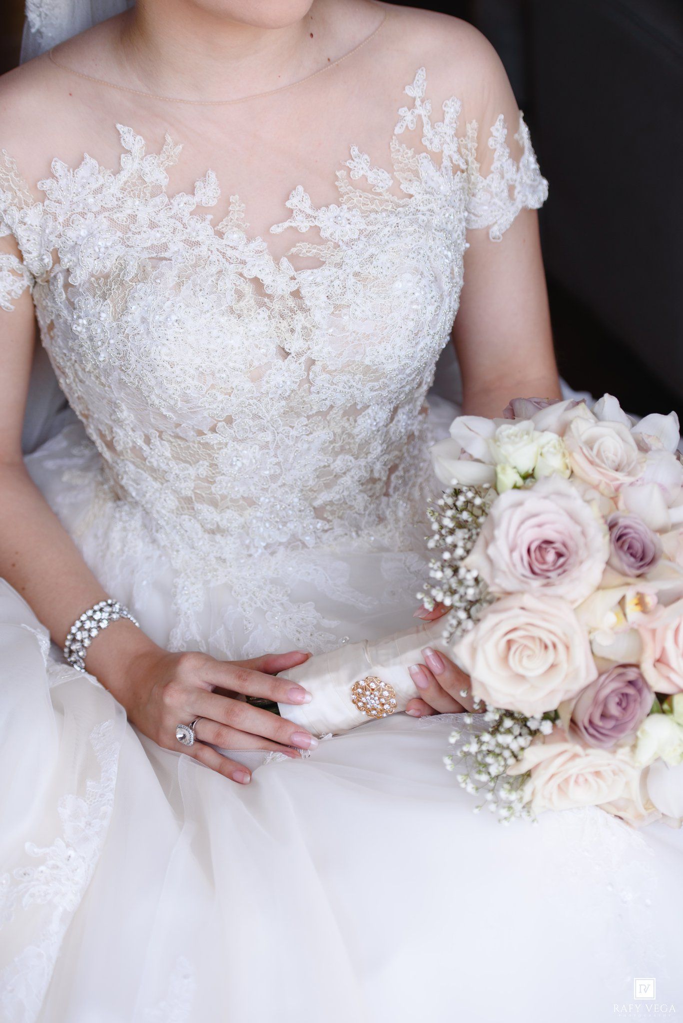 Bride in lace wedding gown holding a bouquet of blush and lavender roses with baby's breath, adorned with jewelry.