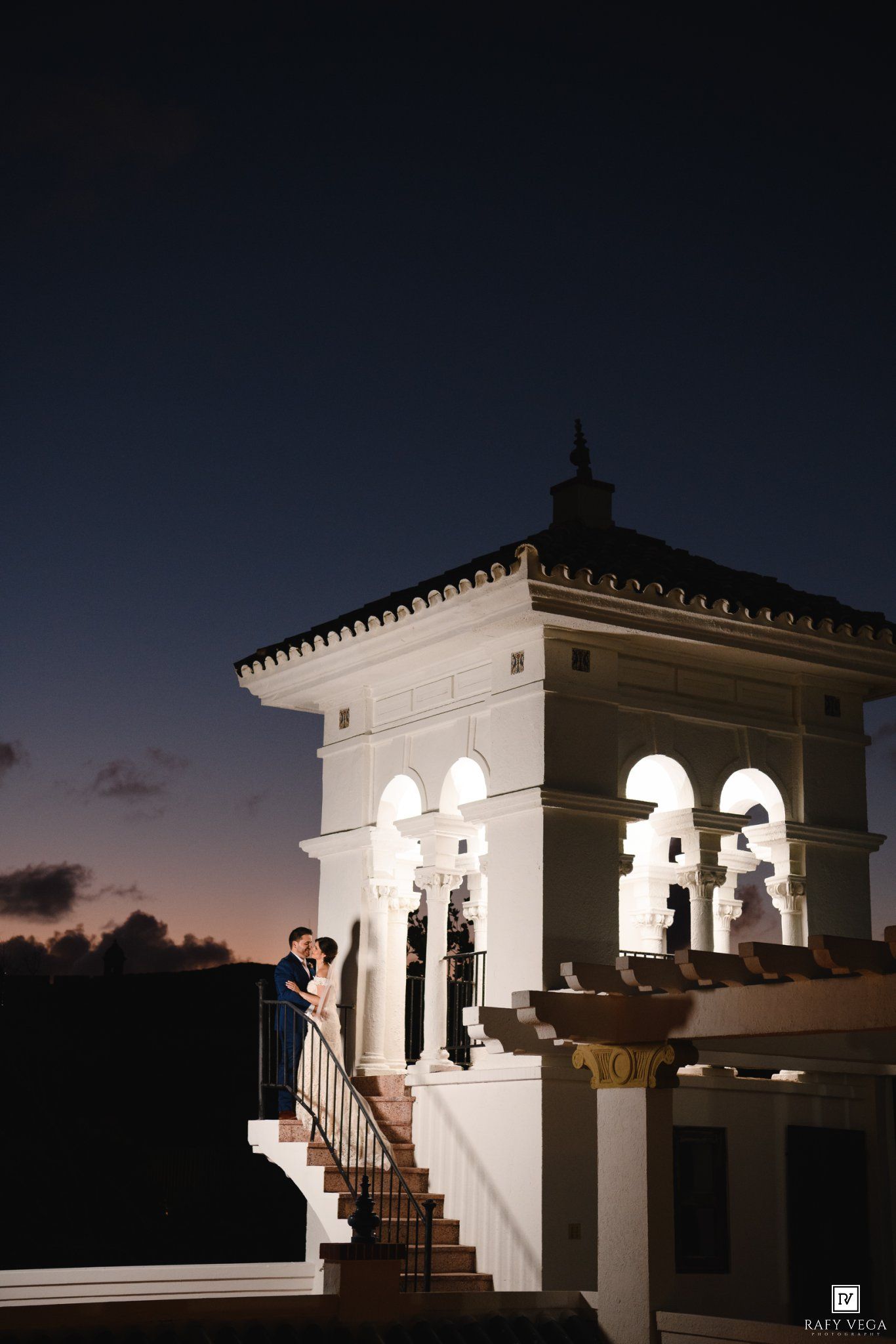 Couple shares romantic moment on illuminated white tower balcony at twilight during elegant outdoor wedding celebration.