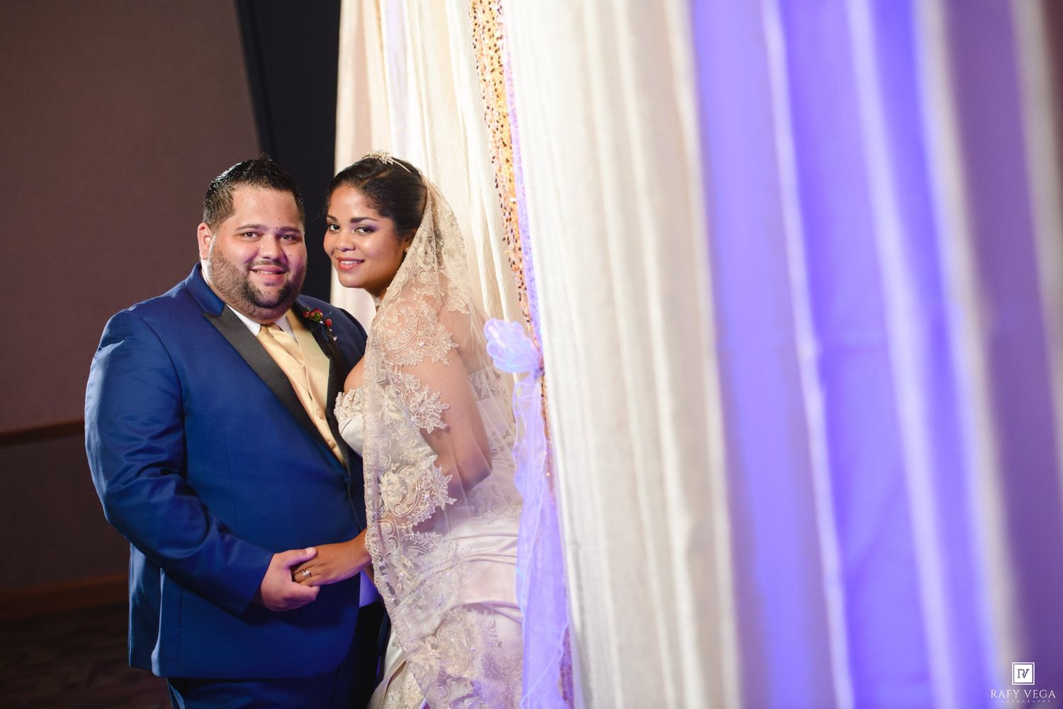 A bride in a lace veil and gown poses with a groom in a blue suit at an elegant wedding reception.