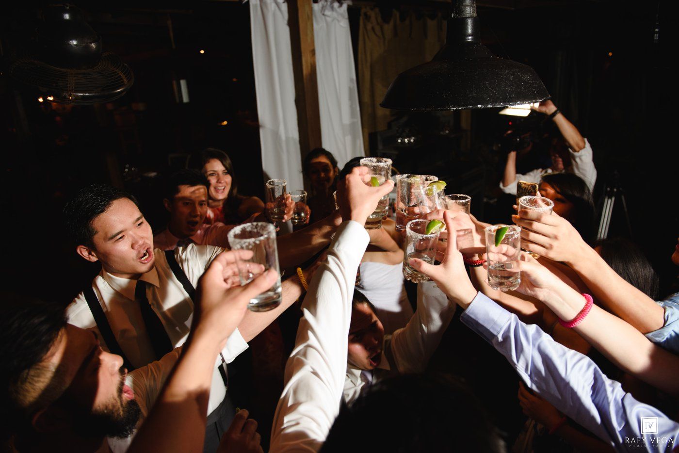 Wedding guests raising glasses in a joyful toast at a dimly lit reception celebration.