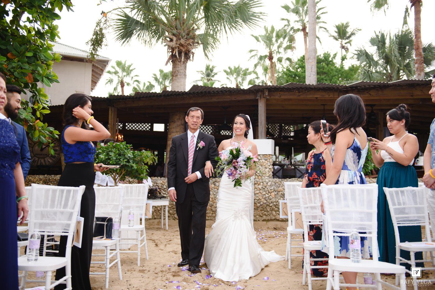 Bride and groom walk down sandy outdoor wedding aisle lined with white chairs and guests in tropical venue.