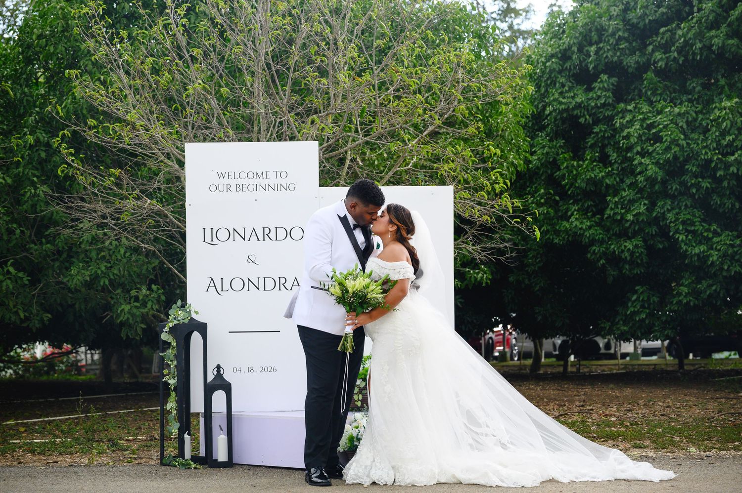 Couple kissing at outdoor wedding ceremony in front of Leonardo and Alondra welcome sign, bride in white gown.