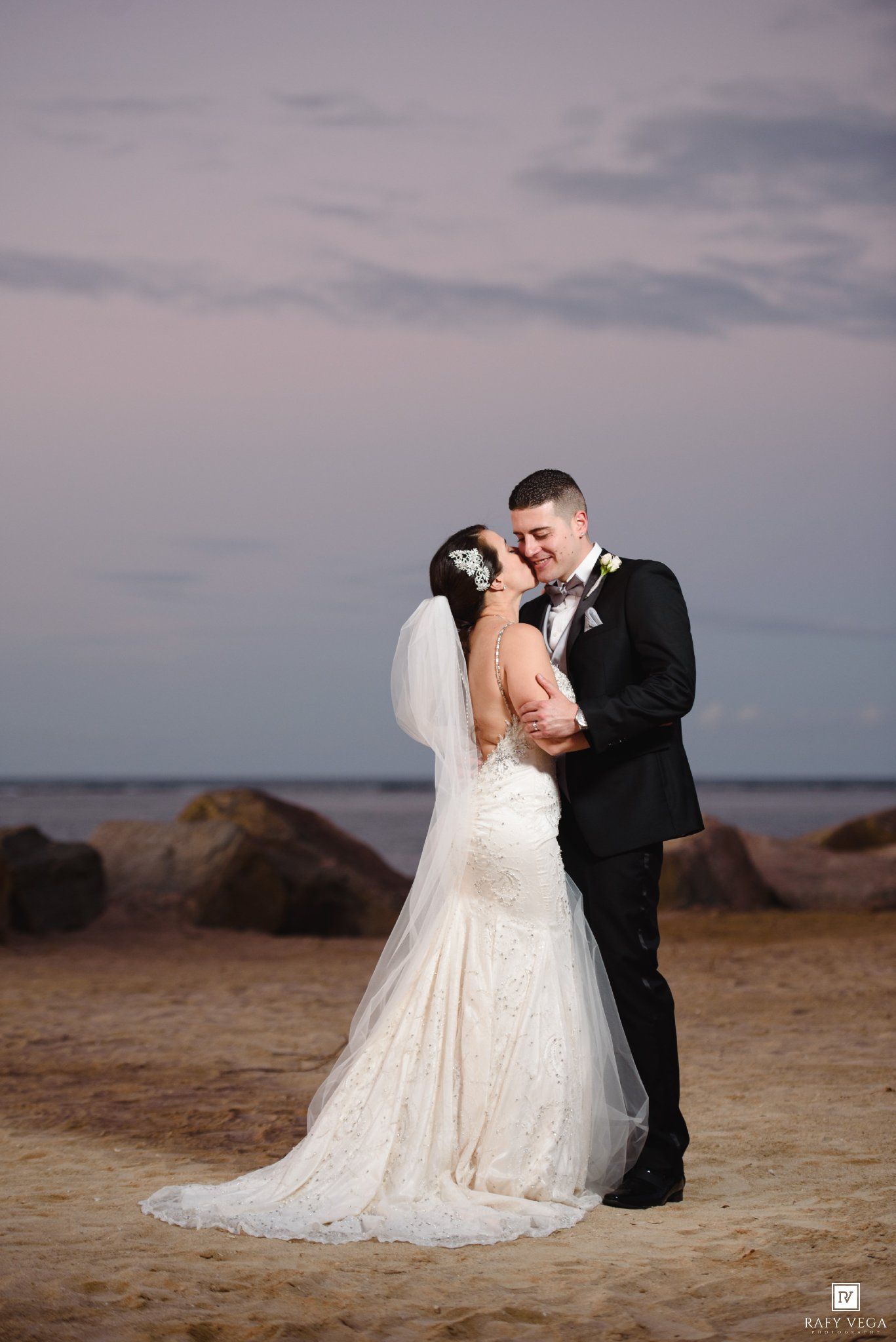 Bride and groom share a romantic kiss on a sandy beach at sunset, bride in lace gown with veil, groom in black tuxedo.