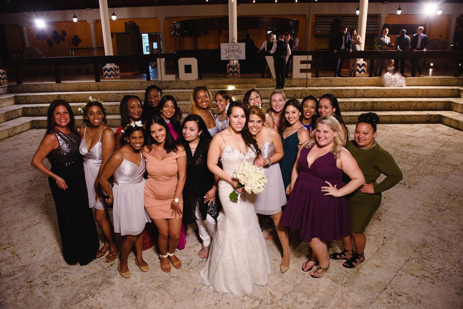 Bride in white gown poses with bridesmaids in colorful dresses at an outdoor evening wedding reception.