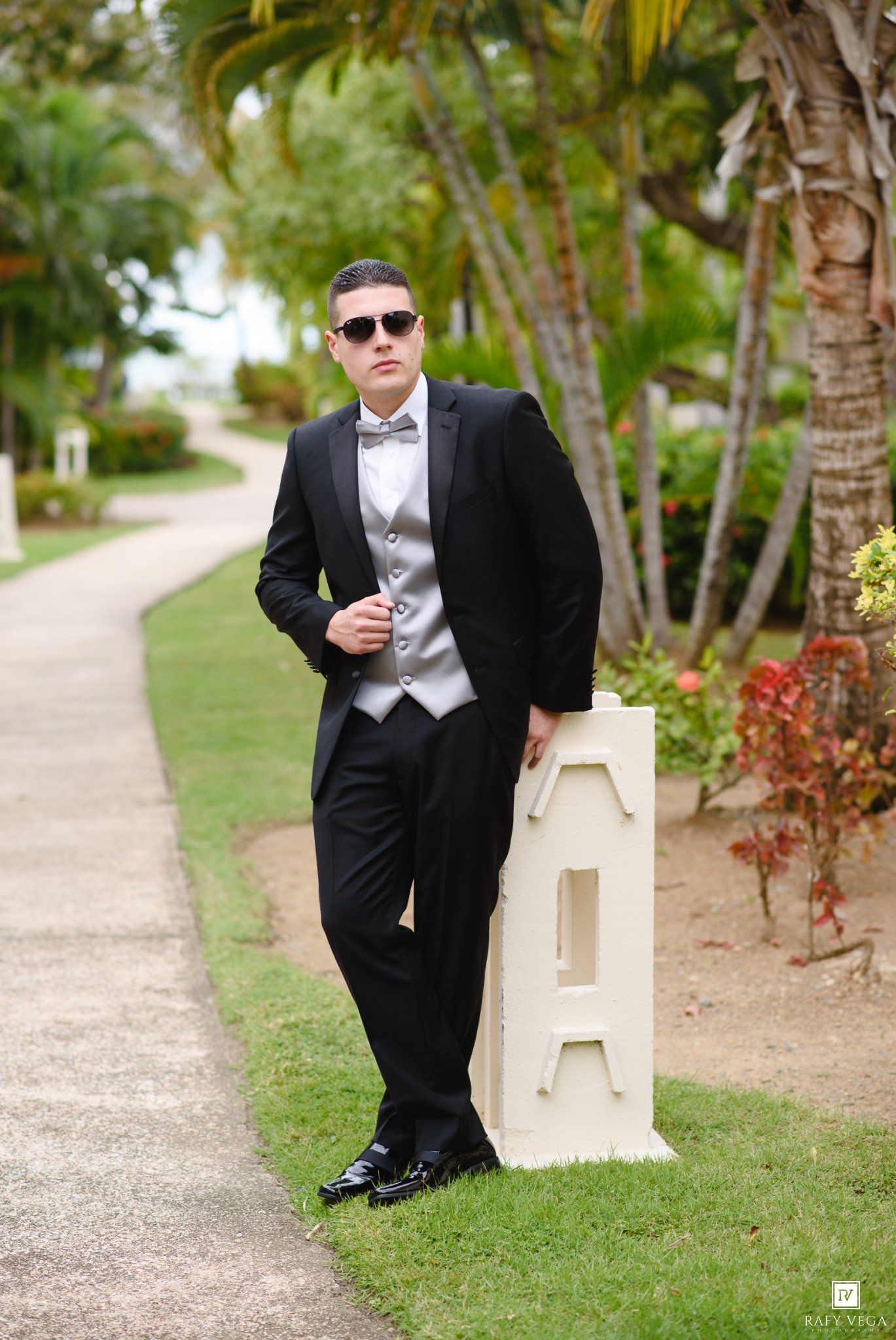 Young man in black tuxedo with gray vest and sunglasses posing outdoors near letter A decoration and palm trees.