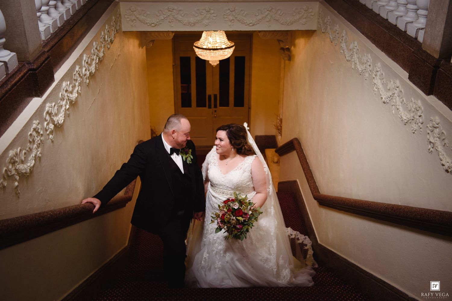 Bride and groom share a romantic moment on an elegant staircase, bride holding a lush bouquet in a grand venue.