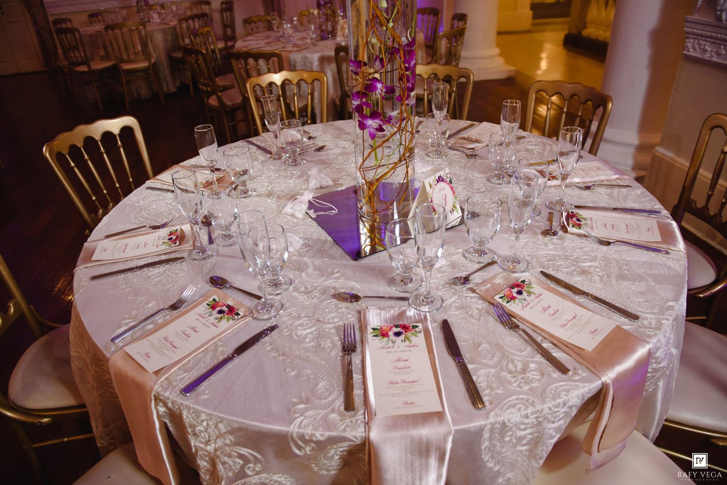 Elegant round banquet table set with white lace tablecloth, crystal glasses, and purple floral centerpiece for a formal event.