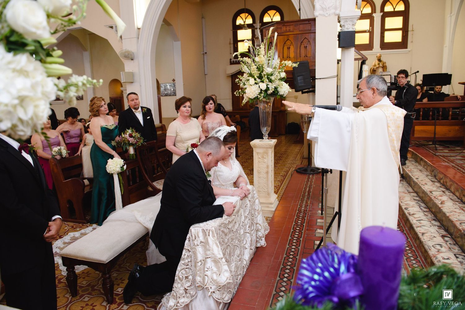 Bride and groom kneeling before priest during Catholic wedding ceremony in ornate church with bridesmaids in background.
