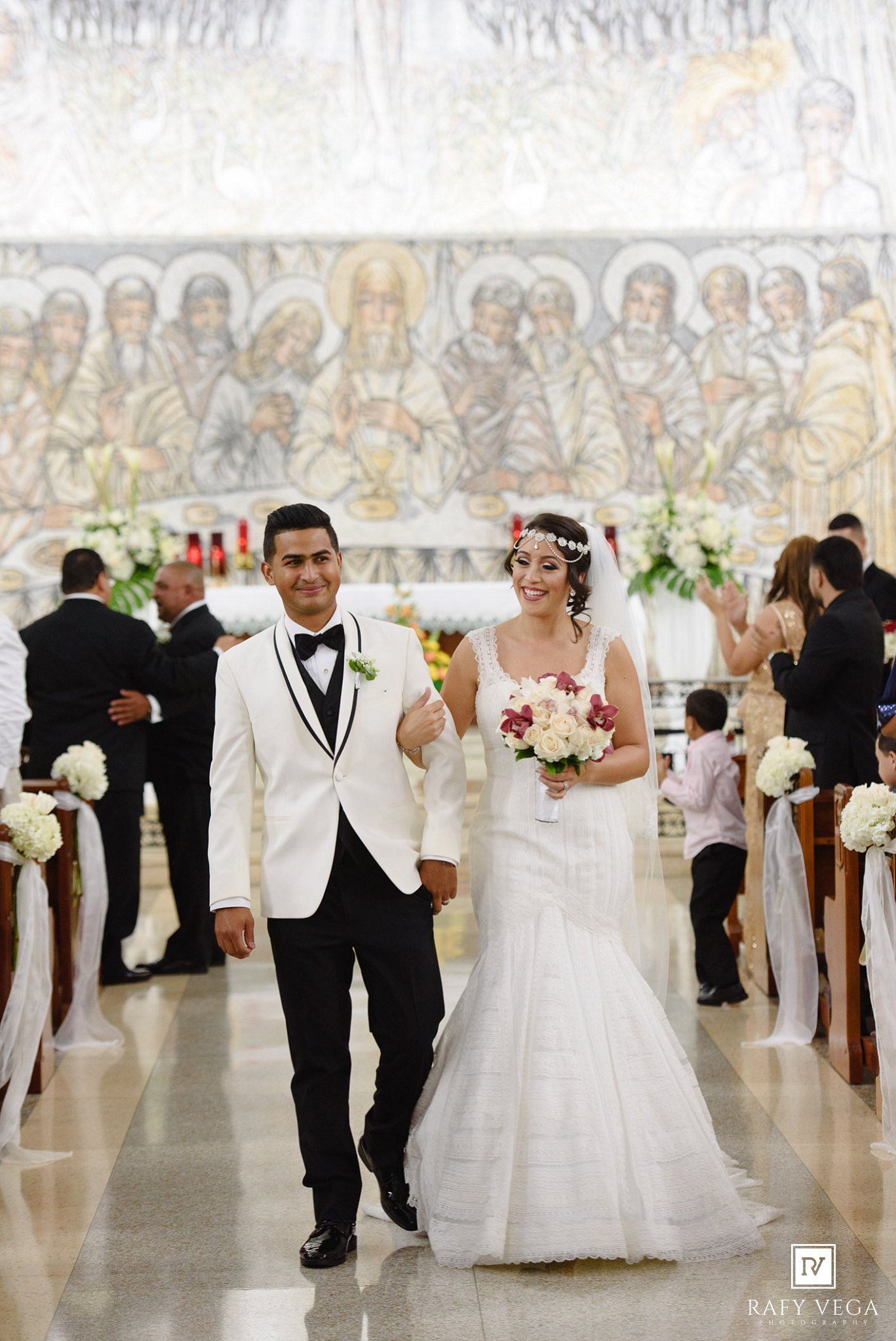 Bride and groom walking down church aisle after wedding ceremony, bride holding bouquet, groom in white tuxedo.