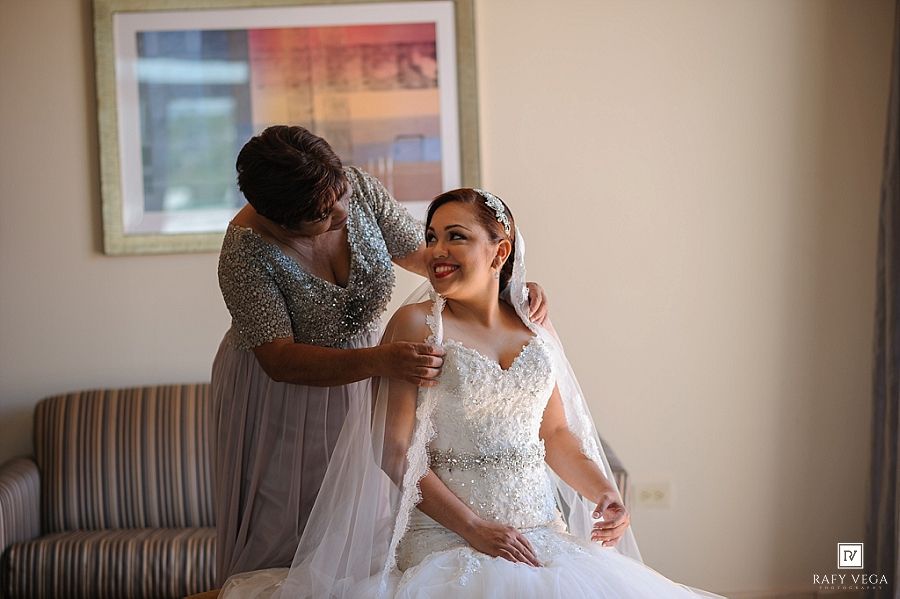 A bride in a white gown smiles as a woman in grey adjusts her veil during wedding preparations indoors.