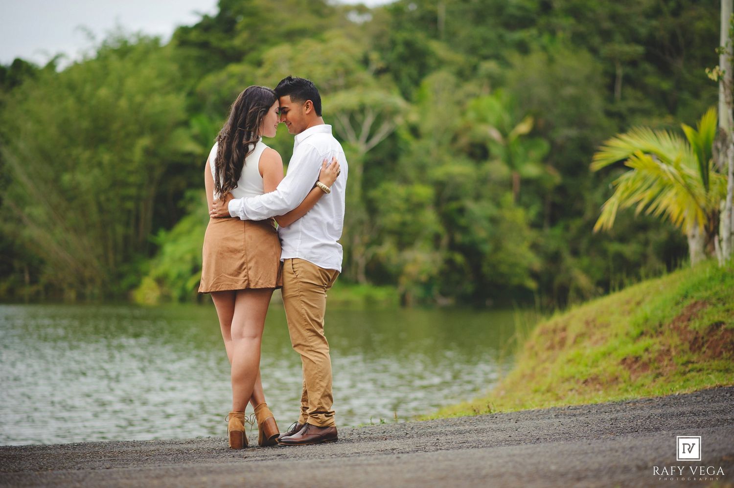 Couple sharing a romantic moment by a serene lake surrounded by lush green tropical trees and palm trees.