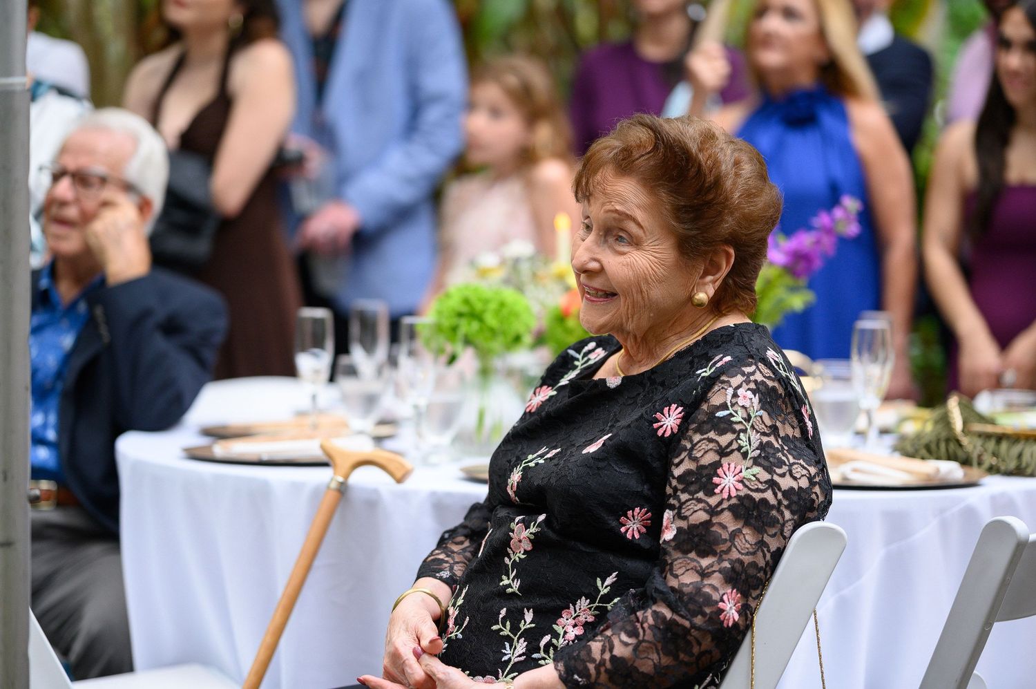 Elderly woman in floral dress smiling at outdoor wedding reception with guests and decorated tables in background.