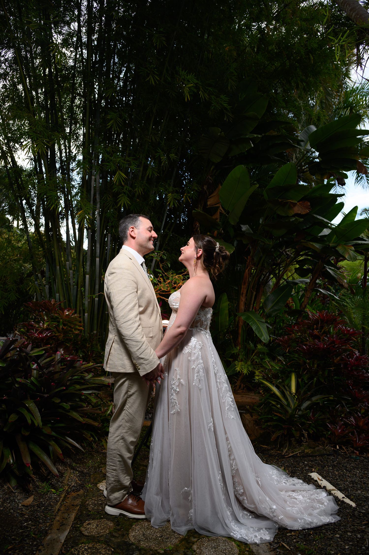 Bride and groom holding hands and smiling at each other in a lush tropical garden setting.